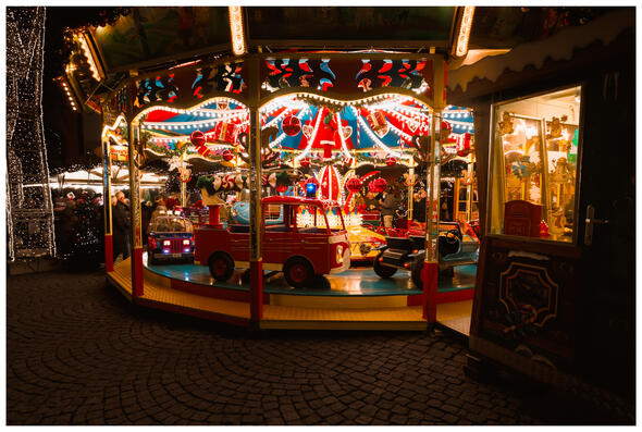 A photograph of an empty, brightly-lit childrens' merry-go-round in vivid colours. In the background, Christmas market visitors are clustered around a drinks stall and standing tables.