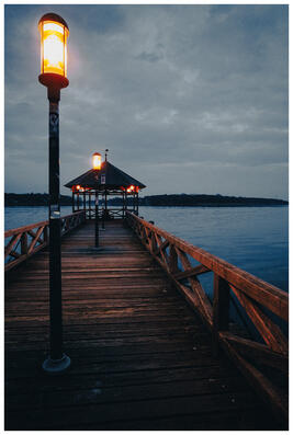 A photograph of a wooden walkway leading onto a lake during blue hour. The walkway is illuminated by retro-looking lamps, and it ends in a gazebo with benches.