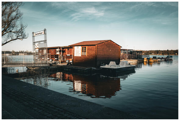 A photograph of a wooden boathouse on a lake. One side of the house is lit by the sun, turning the dark wood vibrant.
