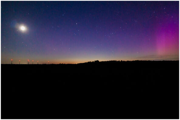 A photograph of the night sky above silhouetted ground. There's a slightly shapeless moon on the left with wind turbines underneath it. The right half of the sky is taken up by a vivid pink aurora.