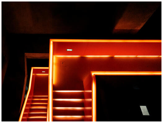 A photograph of layered stairs leading down. The background is black except for a single window reflection at the bottom right of the picture. The orange stairs are illuminated.
