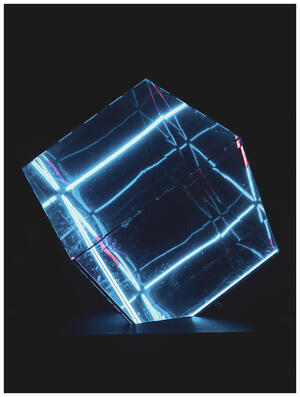 A photograph of a cube made of LED tubes and glass. The tubes are glowing a bright blue with light pink accents. The cube is sitting corners-up with the bottom corner embedded in a platform. The surroundings are dark.