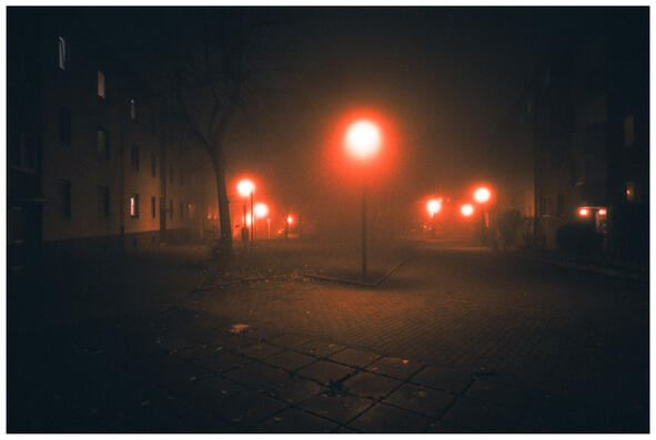 A photograph of a foggy street. The scene is tinged with a reddish hue emanating from the streetlights, which look like glowing orbs.