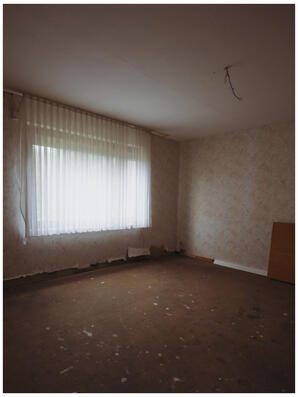 A photograph of a room in an abandoned building. The old patterned wallpaper is peeling up from the bare floor. There is a flimsy curtain hiding the outside from view, diffusing the light.