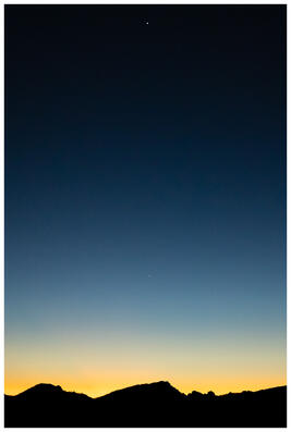 A photograph of a night sky above a silhouetted mountain range. Just above the mountains, there's a hint of fading sunlight.