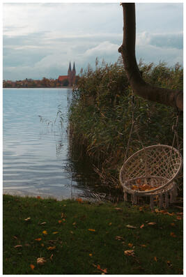 A photograph of a woven chair hanging from a branch by a lakeshore. You can see a church on the opposite shore.