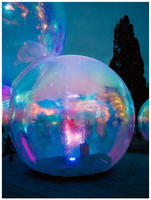 A photograph of a large holo-looking blow-up ball, light in blue and pink. There are two silhouettes in the centre of the ball.